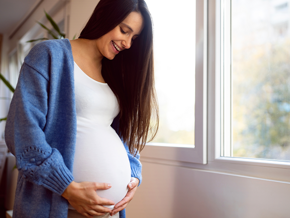 Mujer embarazada vestida de blanco, sonriendo y sosteniendo su vientre con las manos.