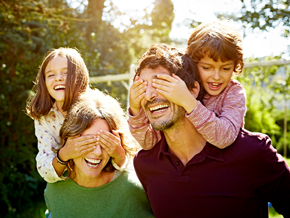 Familia de cuatro disfrutando de un día soleado al aire libre. Dos adultos llevan a dos niños sobre sus hombros, rodeados de árboles y vegetación