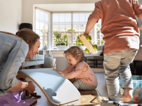 Mujer adulta y dos niños juegan en una sala, con un tablero de equilibrio y un coche de juguete.