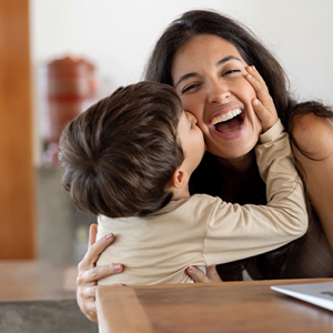 Establecer límites saludables Niño dando un beso en el cachete a su madre sonriente. La mamá está sentada en frente de un ordenador portátil.