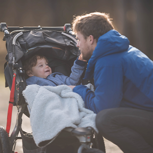Edad adecuada para llevar a tu bebé en cochecito Papá arrodillado junto a un cochecito con un niño cubierto por una manta, en un camino al aire libre.