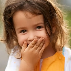 Niña pequeña al aire libre riendo y tapando su boca con su mano.