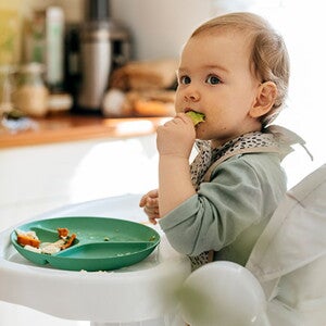 Niño pequeño en silla alta comiendo verduras con las manos.