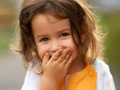 Niña pequeña al aire libre riendo y tapando su boca con su mano.