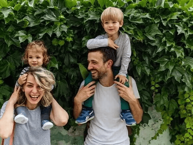 Dos adultos con dos niños sobre sus hombros, frente a una pared cubierta de hojas. 