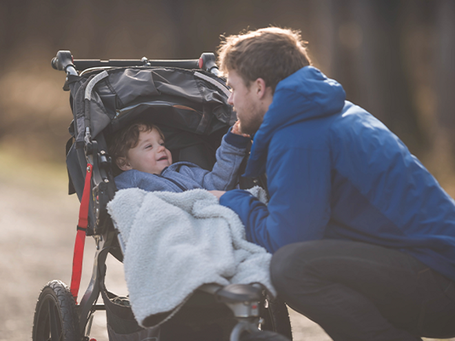 Edad adecuada para llevar a tu bebé en cochecito Papá arrodillado junto a un cochecito con un niño cubierto por una manta, en un camino al aire libre.
