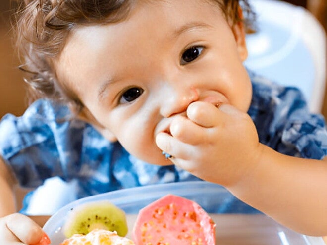 Niño pequeño sentado en silla alta comiendo frutas con las manos.