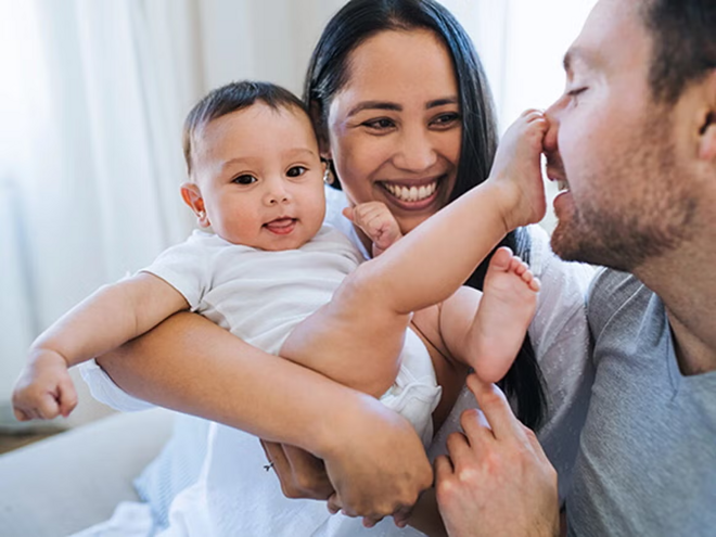 ¿Criar en equipo? Sí, se puede: descubre los beneficios de la crianza compartida Mamá carga al bebé sonriendo. Bebé tiene el pie en la nariz del papá.