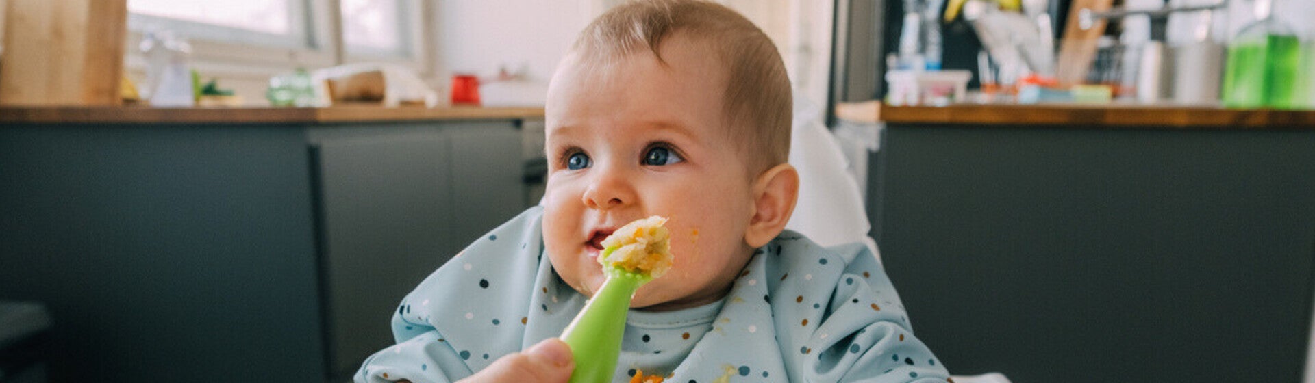 Mano de mamá dando de comer con una cuchara a su bebé que está sentado en silla alta. Alimentación complementaria del bebé.