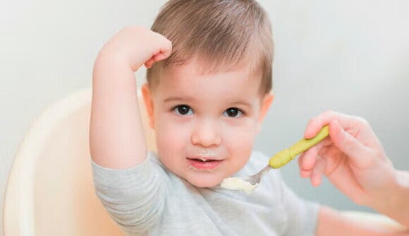 Niño sonriendo a la camara mientras su mamá le da de comer papilla Niño sonriendo a la camara mientras su mamá le da de comer papilla